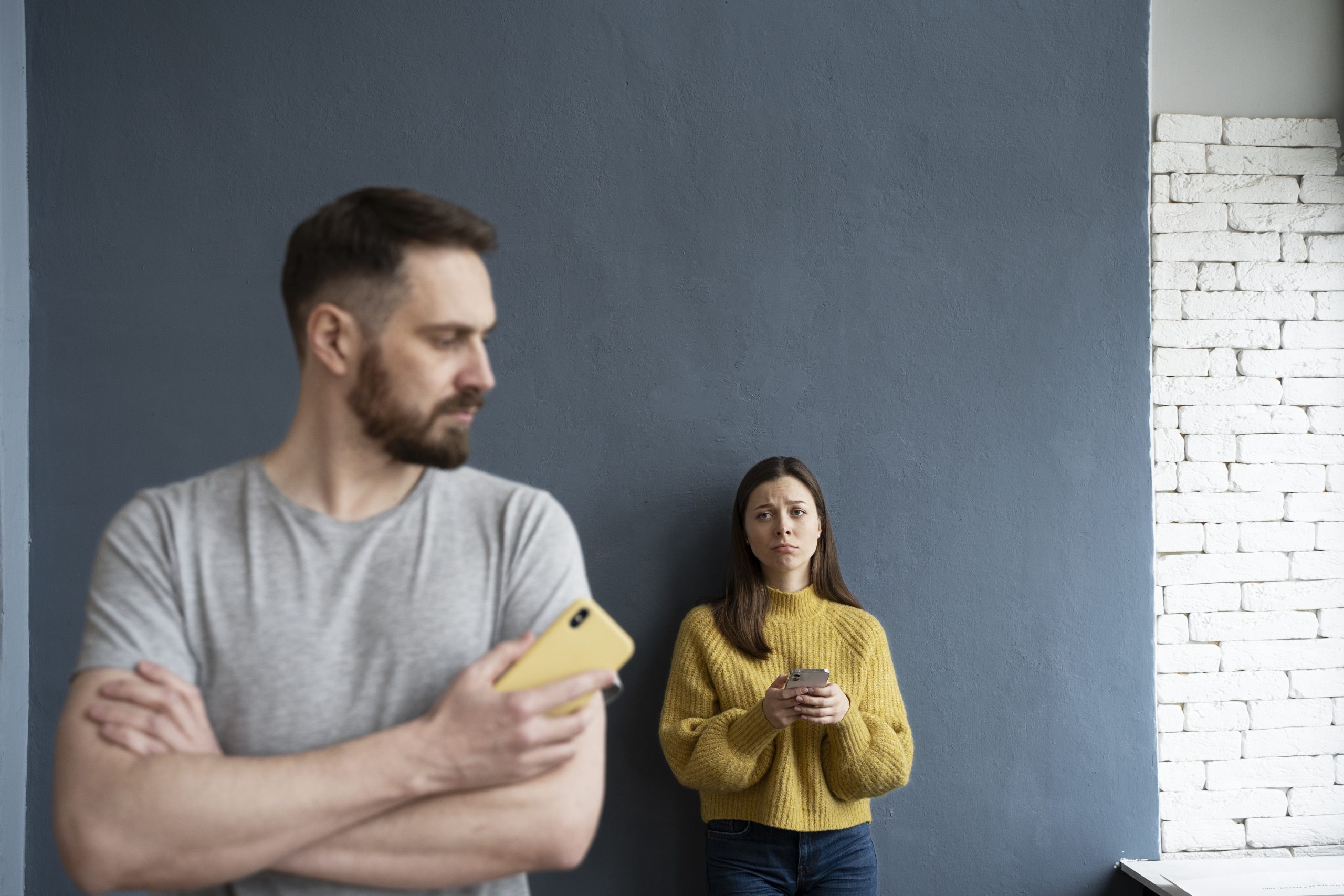 Homem de braços cruzados segurando um celular amarelo e olhando para o lado, enquanto uma mulher ao fundo, usando um suéter amarelo, segura um celular e aparenta estar triste ou preocupada