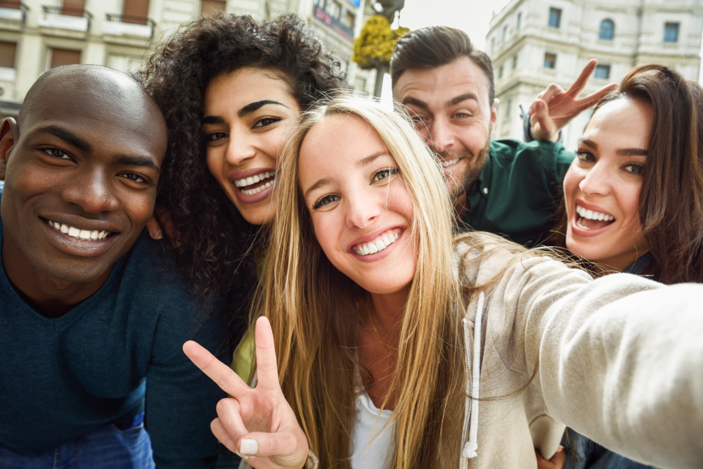 Grupo de cinco amigos sorridentes tirando uma selfie ao ar livre em um ambiente urbano. Todos estão próximos uns dos outros, demonstrando alegria e descontração