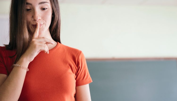 Mulher com blusa laranja e cabelos pretos com dedo na boca sugerindo silêncio