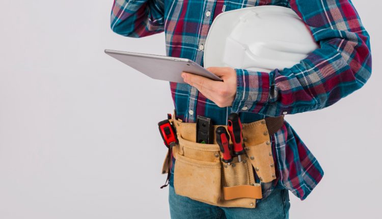 Trabalhador técnico ou engenheiro segurando um tablet e um capacete branco, vestindo camisa xadrez e cinto de ferramentas com chaves de fenda e fita métrica