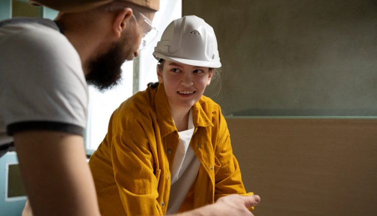 Dois profissionais conversando em um ambiente de construção. Uma mulher usa capacete branco e jaqueta amarela, sorrindo enquanto interage com um colega de trabalho que veste boné e óculos de segurança