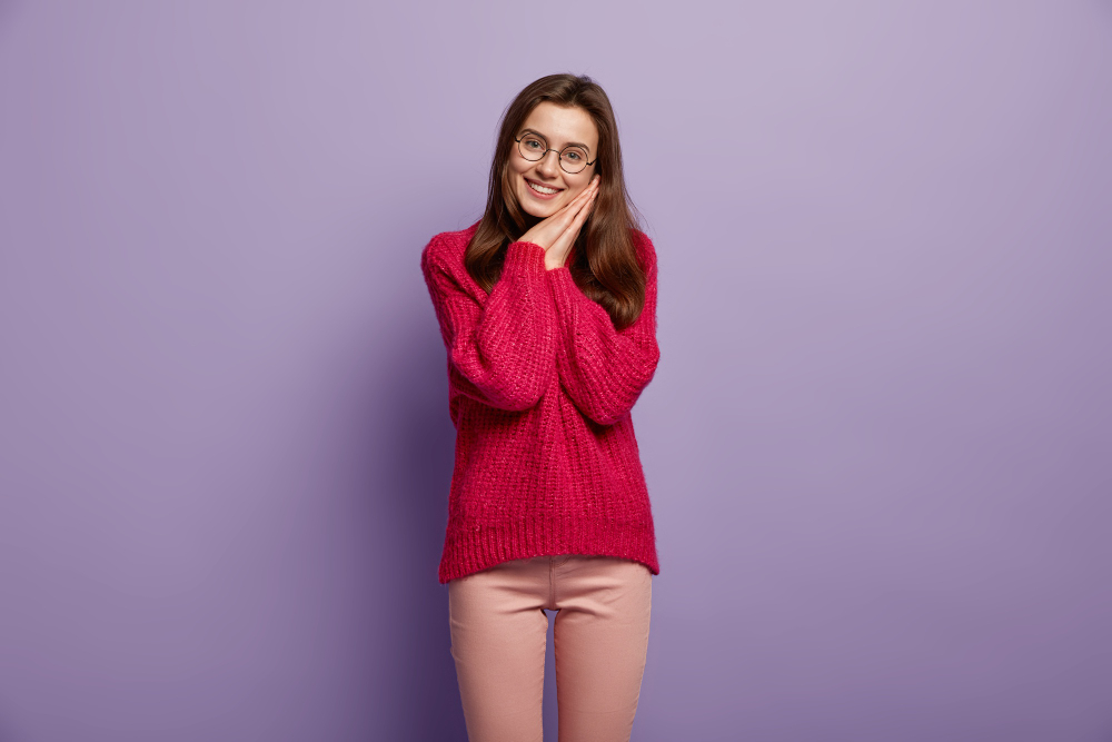 Mulher jovem com óculos e cabelos longos, sorrindo e apoiando o rosto nas mãos. Ela veste um suéter rosa e calça clara, posando contra um fundo roxo