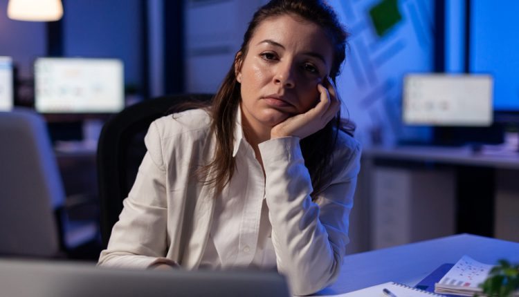 Mulher exausta no escritório, vestindo roupa formal, apoiando o rosto na mão com expressão de cansaço