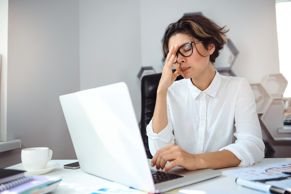 Mulher com expressão de cansaço e estresse no escritório, usando óculos e roupa social branca. Ela massageia a testa enquanto trabalha no laptop, sugerindo fadiga profissional ou burnout