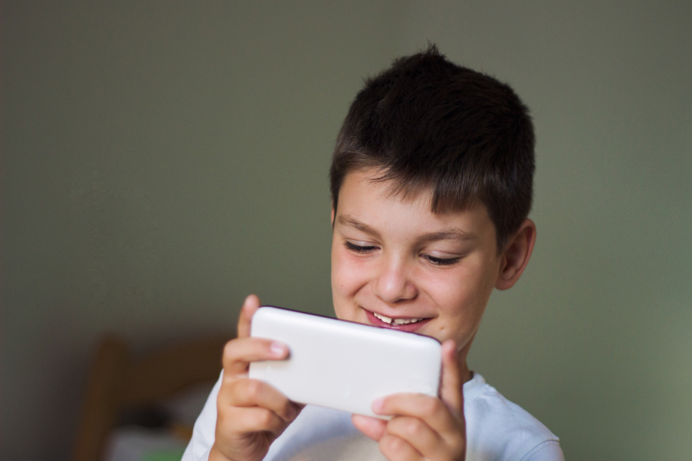 Menino sorrindo se divertindo com o celular na mão.