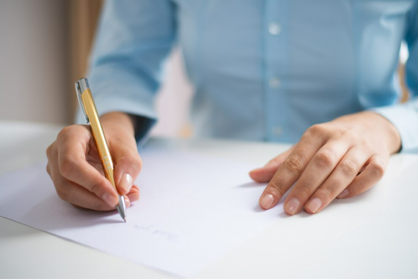 Pessoa escrevendo em uma folha de papel com uma caneta dourada, vestindo uma camisa azul, em um ambiente de escritório