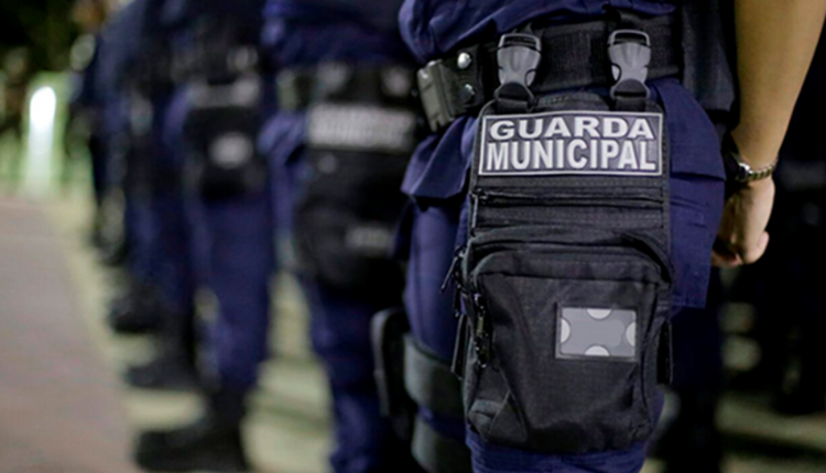 Grupo de agentes da Guarda Municipal enfileirados, vestidos com uniformes azul-escuros e equipamentos táticos. Em destaque, uma bolsa preta presa à perna de um dos agentes com o nome 'GUARDA MUNICIPAL' em destaque