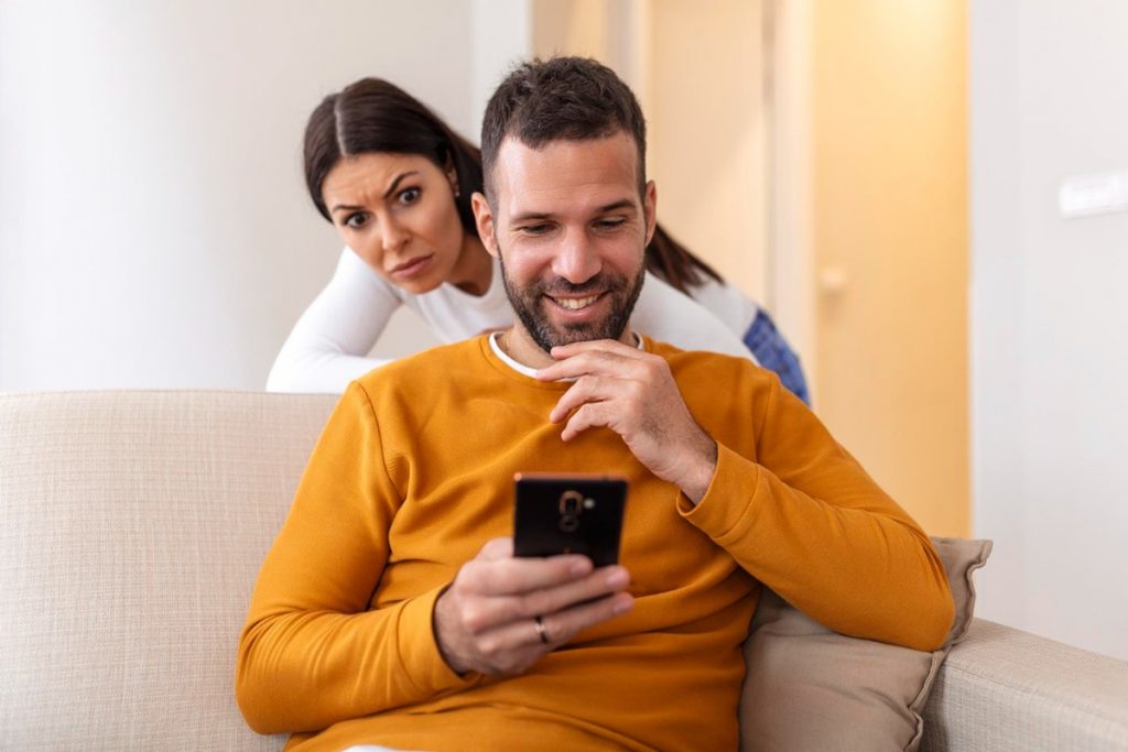 Mulher observando com desconfiança um homem sorrindo enquanto usa o celular.