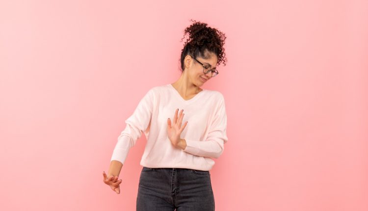 Mulher de cabelos cacheados e óculos, vestindo blusa clara e calça jeans, fazendo um gesto de recusa com as mãos, expressando desconforto, contra um fundo rosa