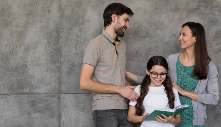 Menina com óculos e cabelo preso em tranças lendo um livro, sorrindo ao lado de um casal que a observa com carinho, contra um fundo de parede cinza.