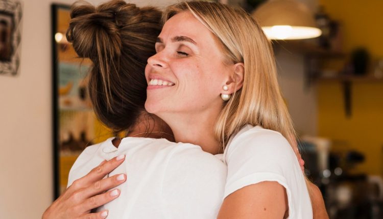 Duas mulheres sorrindo e se abraçando, com um momento de conexão e amizade, em um ambiente acolhedor