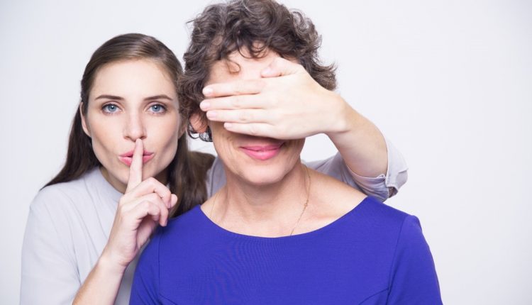 Mulher fazendo gesto de silêncio enquanto cobre os olhos de outra pessoa