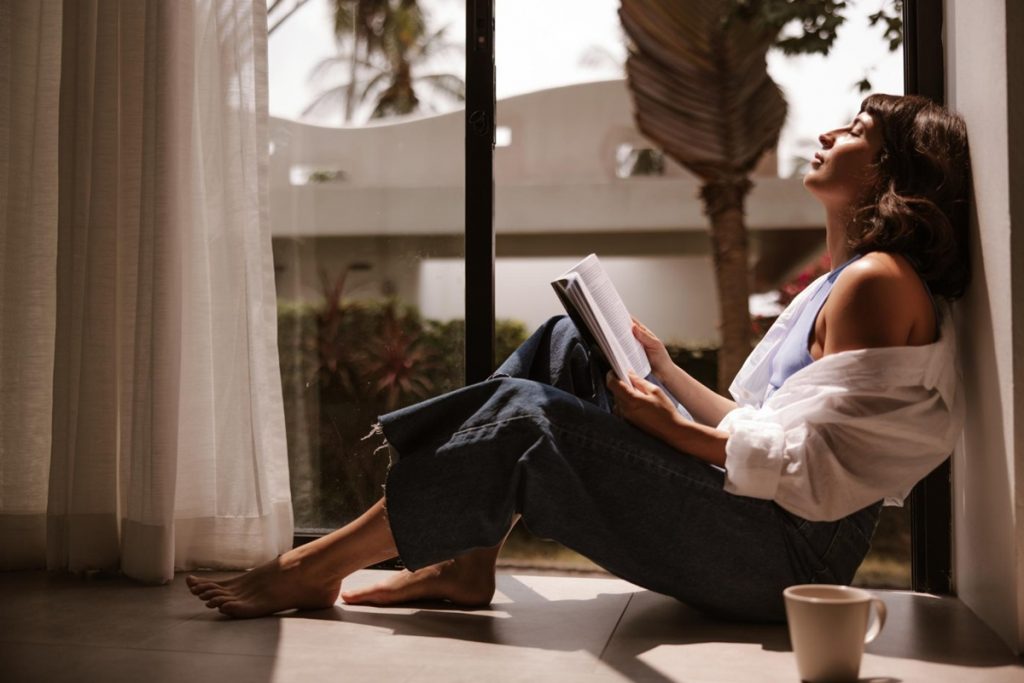 Mulher relaxando enquanto lê um livro próximo à janela, aproveitando a luz natural