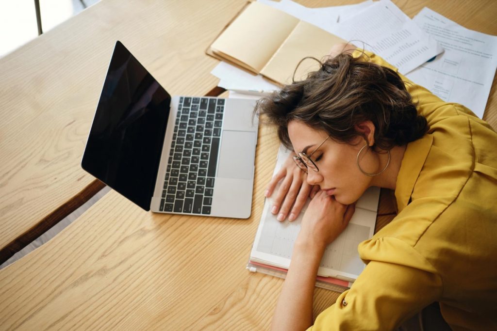 Mulher exausta dormindo sobre a mesa com o laptop aberto e papéis espalhados, representando os efeitos da procrastinação crônica.