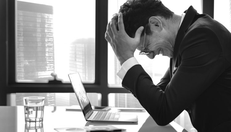 Homem estressado no escritório com as mãos na cabeça, aparentando frustração diante do laptop.