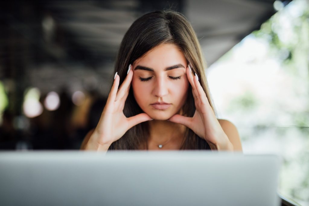 Mulher jovem concentrada com os olhos fechados e as mãos na cabeça, buscando se acalmar diante do laptop