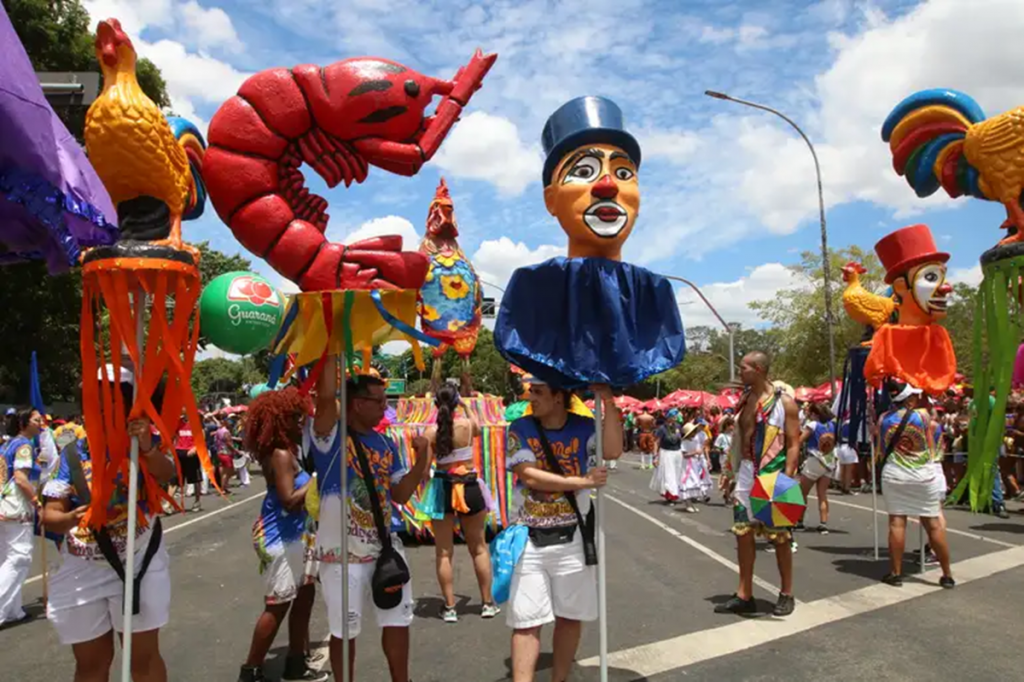 Desfile de carnaval com bonecos gigantes e foliões, representando as festividades do Carnaval 2025.