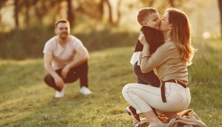 Mãe beijando seu filho no rosto enquanto o pai observa ao fundo em ambiente ao ar livre.