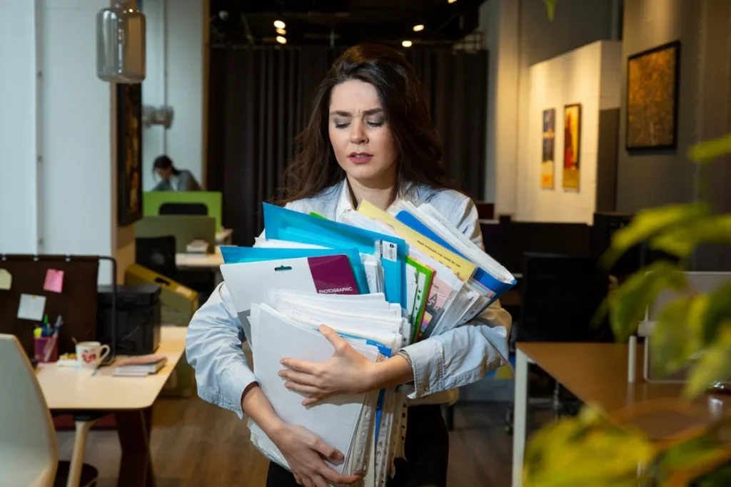 Mulher em escritório carregando pilhas de documentos, ilustrando o impacto da sobrecarga no trabalho e vida pessoal.