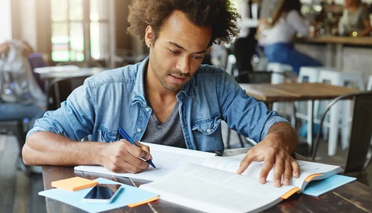 Jovem estudando em um café, concentrado na leitura de um livro e fazendo anotações