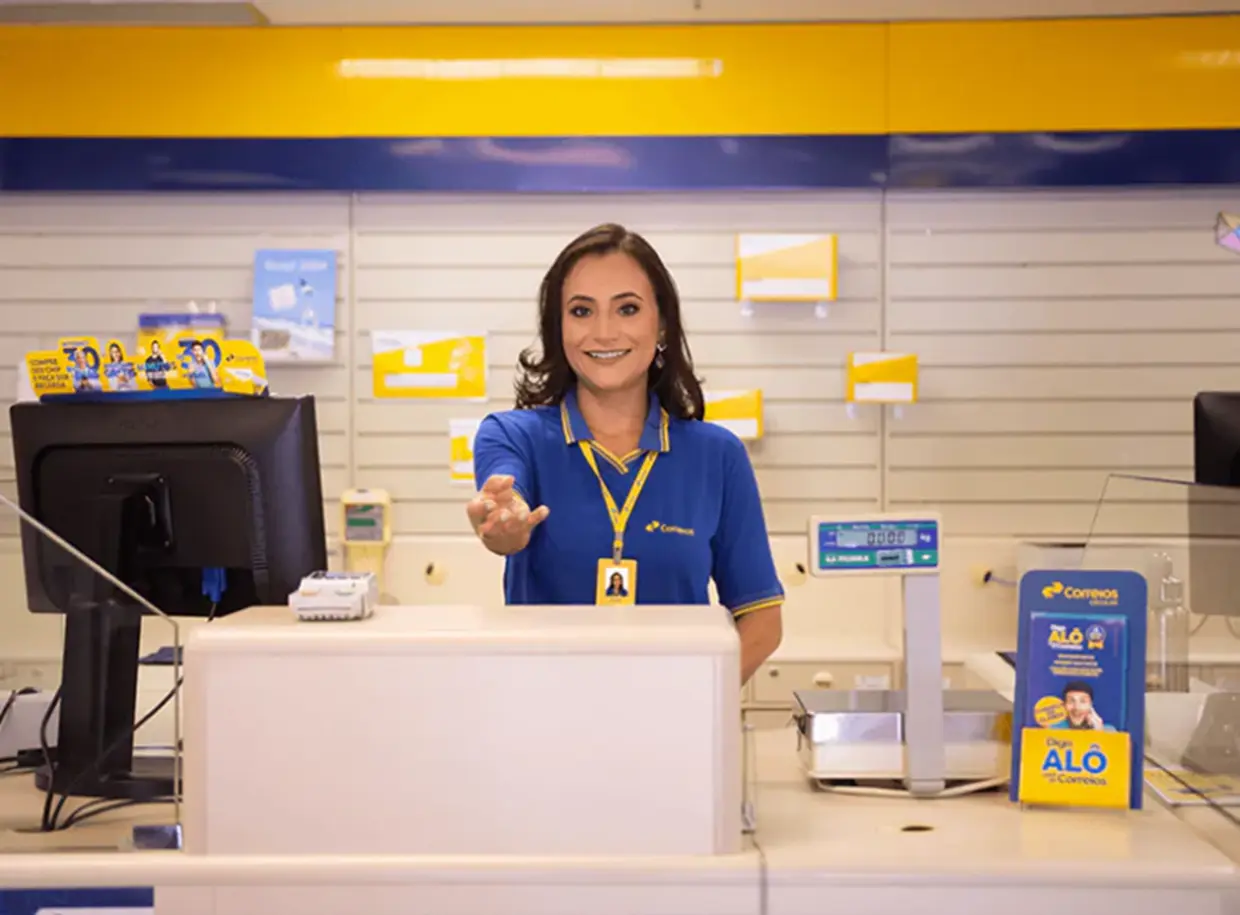 Atendente comercial dos Correios atendendo no balcão de uma agência, com uniforme oficial e sorrindo para os clientes