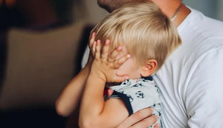 Criança cobrindo o rosto com as mãos enquanto é abraçada, simbolizando dificuldades emocionais na infância.