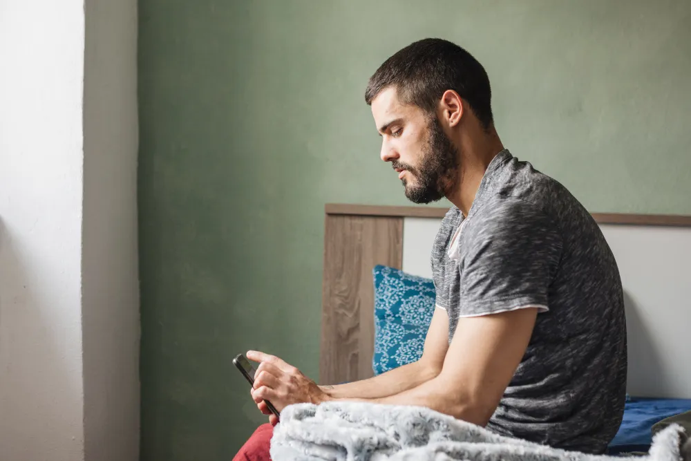 Homem sentado na cama olhando para o celular com expressão reflexiva e preocupada.