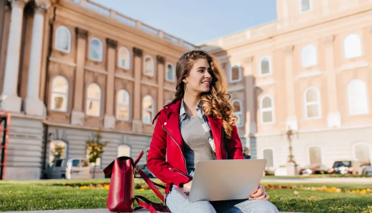 Jovem sorridente com jaqueta vermelha usando um laptop ao ar livre em um ambiente universitário.