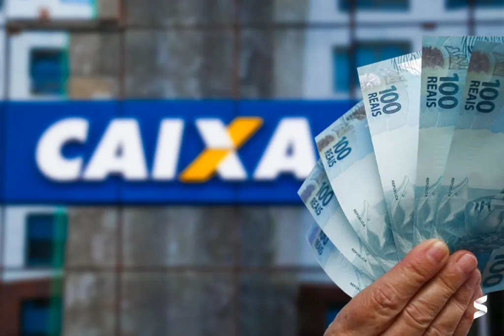 Mão segurando cédulas de R$ 100 em frente a uma agência da Caixa Econômica Federal.