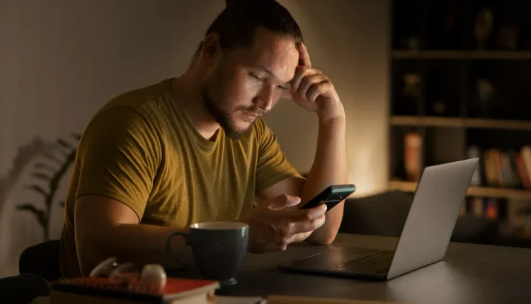 Homem pensativo segurando um celular enquanto trabalha à noite, refletindo sobre seus hábitos diários.