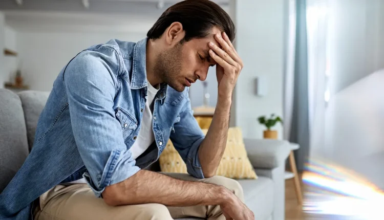 Homem jovem sentado no sofá, segurando a cabeça com expressão de esquecimento