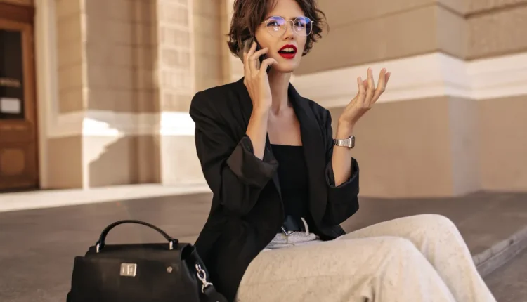 Mulher com cabelo curto, óculos e batom vermelho fala ao celular enquanto gesticula, sentada ao ar livre com uma bolsa ao lado.