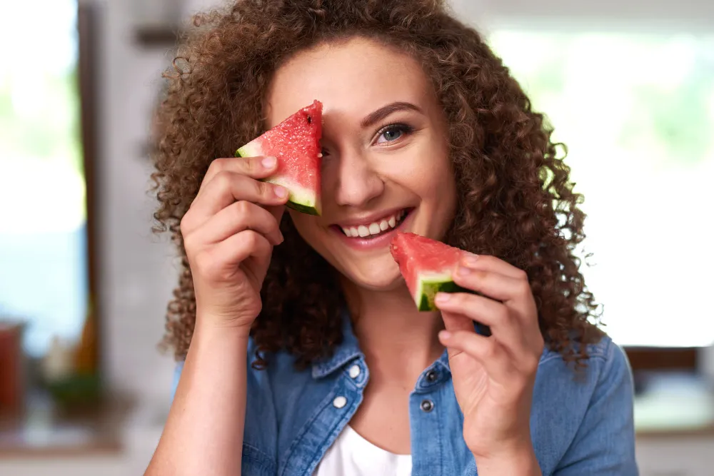 Mulher sorrindo e segurando pedaços de melancia, representando alimentação saudável e refrescante.