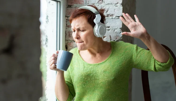 Mulher ouvindo música com fones de ouvido enquanto toma café, sorrindo e se movendo ao ritmo da canção, representando a relação entre música e emoções.