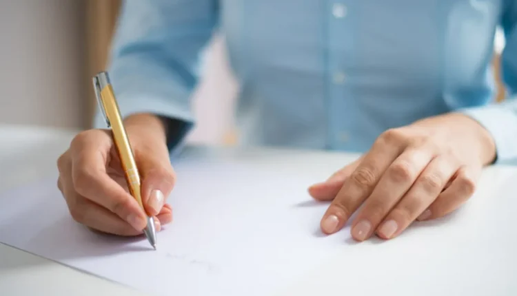 Mão escrevendo em uma folha em ambiente de escritório, representando a preparação para o concurso público.