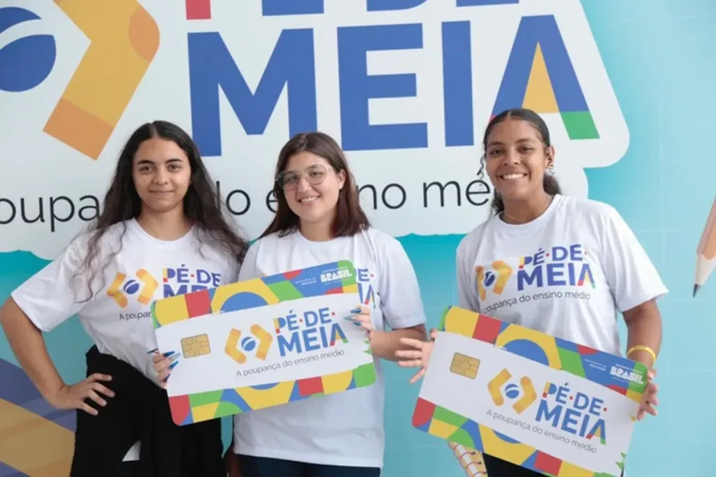 Três estudantes segurando cartões gigantes do programa Pé-de-Meia, simbolizando a poupança do ensino médio.