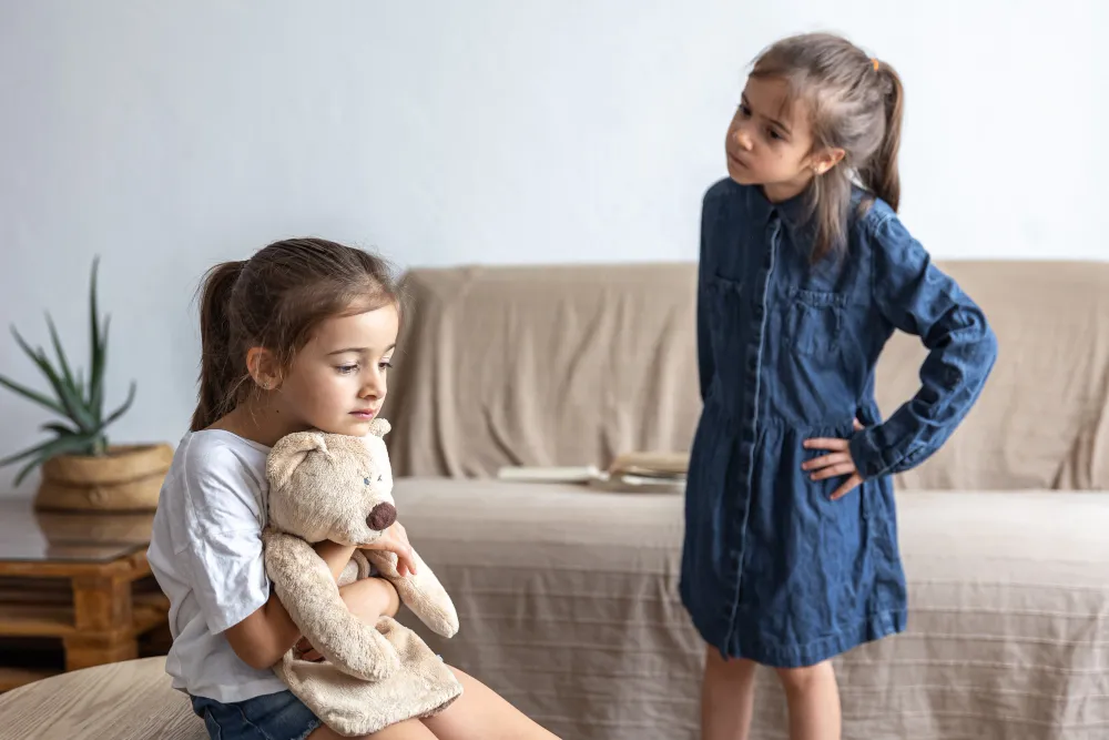 Duas meninas, uma com um ursinho e a outra observando, simbolizando a privação emocional na infância.