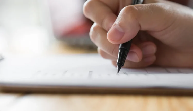 Mão segurando uma caneta e preenchendo um formulário, simbolizando inscrições em processos seletivos