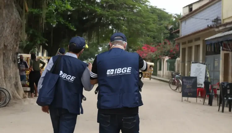 Dois agentes do IBGE caminham em uma rua de terra, vestidos com coletes e bonés oficiais, carregando bolsas institucionais.