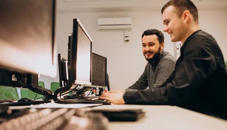 Dois profissionais em um ambiente de escritório, trabalhando juntos em frente a computadores, sorrindo enquanto colaboram