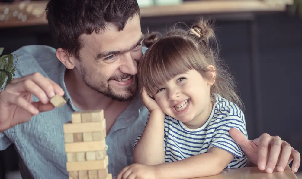 Pai e filha sorrindo juntos enquanto brincam de construir uma torre de blocos, ilustrando o vínculo familiar.