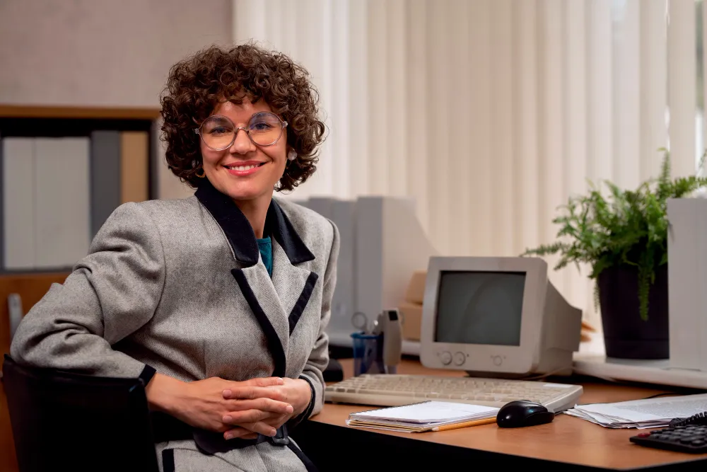 Profissional de secretariado em ambiente corporativo, sorrindo e com um computador à sua frente, representando a organização e eficiência no escritório