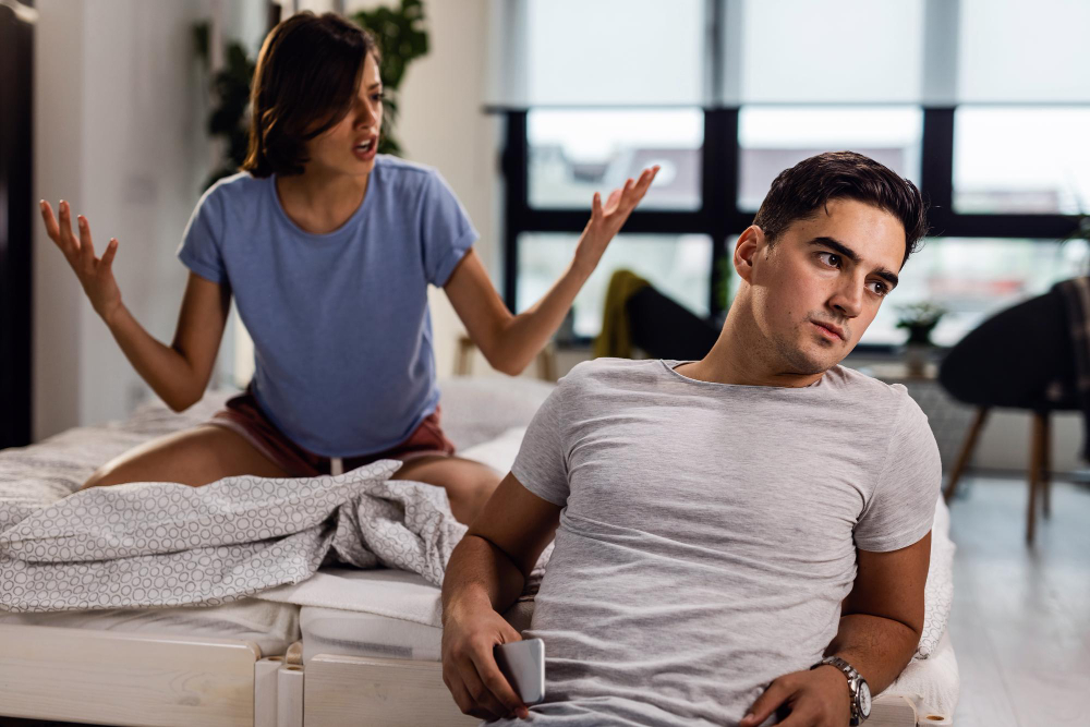 Mulher irritada gesticulando enquanto um homem desvia o olhar com indiferença, durante uma discussão no quarto.