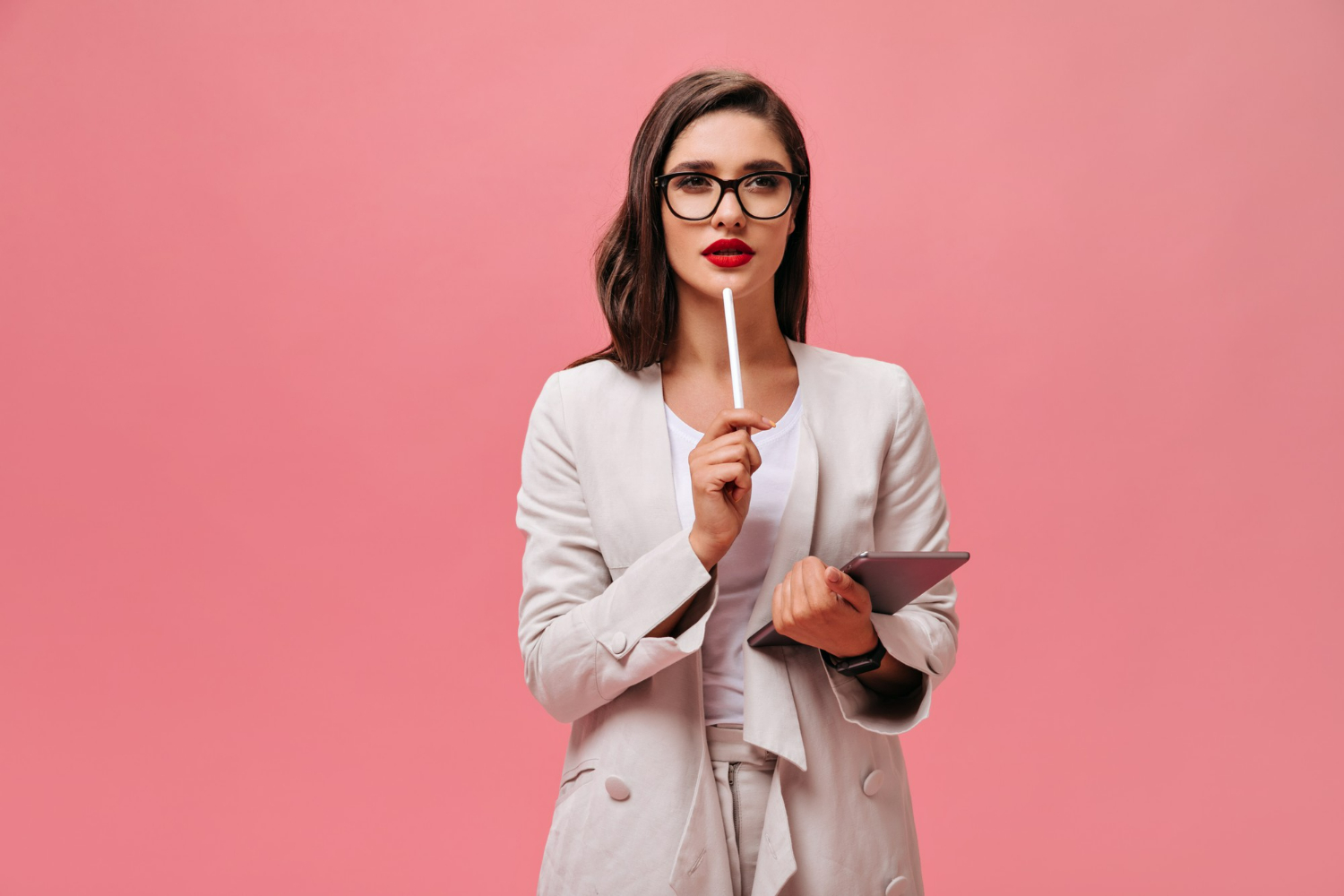 Mulher com óculos e blazer branco, pensativa, segurando uma caneta e um tablet, refletindo sobre perfeccionismo.