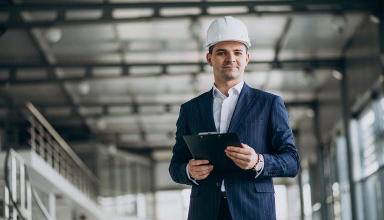 Engenheiro civil usando capacete e segurando uma prancheta em ambiente industrial.