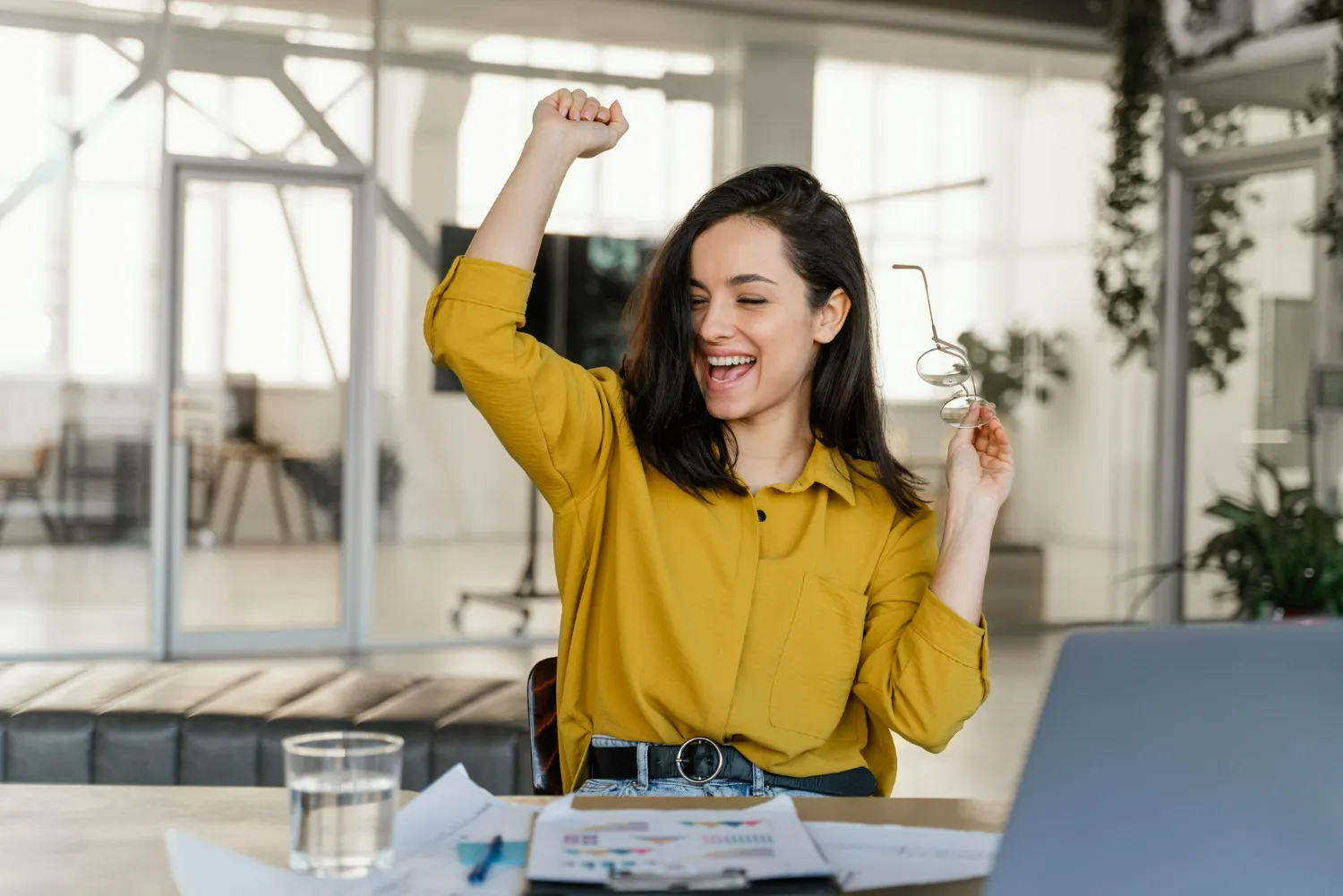 Mulher sorridente levantando o braço em comemoração, com óculos na mão, simbolizando sucesso e realização na profissão de vendas.