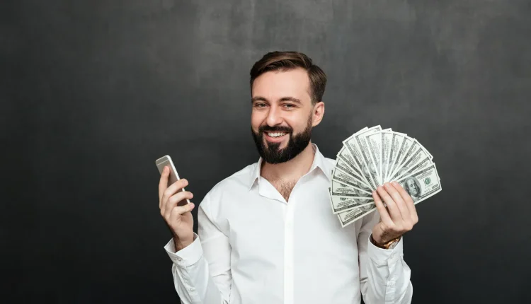 Homem sorrindo e segurando notas de dólar em uma mão e um celular na outra, simbolizando sucesso financeiro.