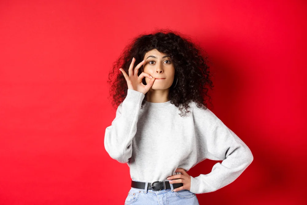 Mulher com expressão pensativa, tocando o rosto e fazendo um gesto de 'silêncio', simbolizando reflexão e autocontrole verbal.