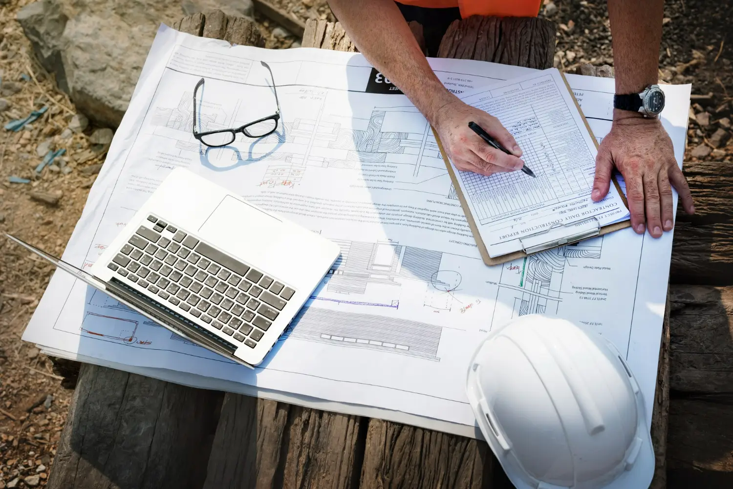 Profissional de engenharia analisando planos de construção com laptop e documentos sobre a mesa.