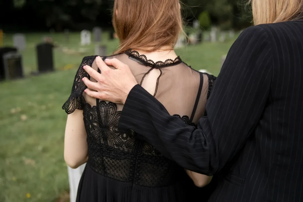 Mulher sendo consolada durante o luto, com um gesto de apoio ao fundo do cemitério.